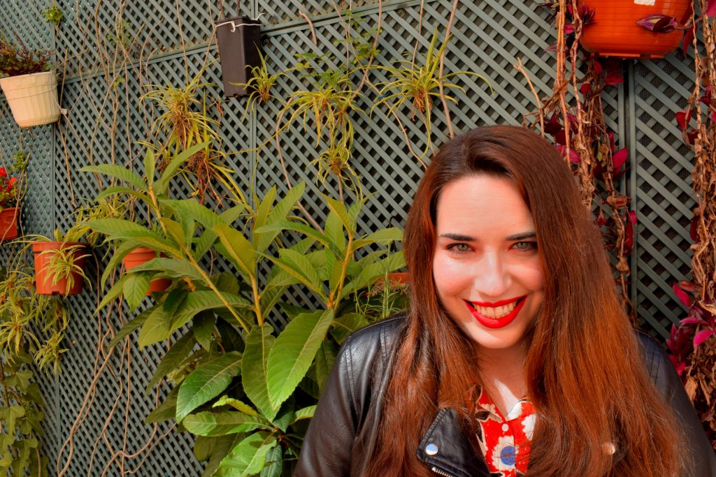 Foto mía en la que aparezco sonriente con los labios rojos en un jardín delante de varias macetas.
